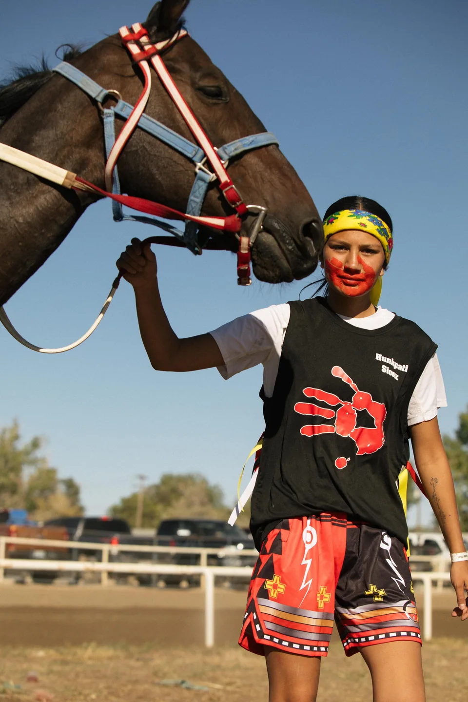 A rider in a black shirt and red shorts stands beside a horse at the racing grounds.