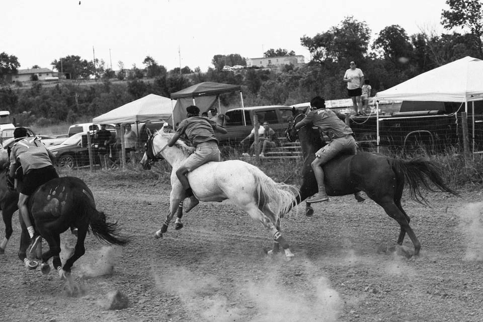 Black-and-white photograph of bareback riders racing past tents and spectators.