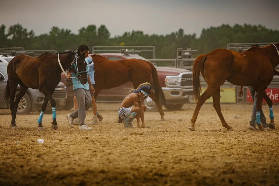 Riders and horse holders prepare between horses on the dusty relay track.