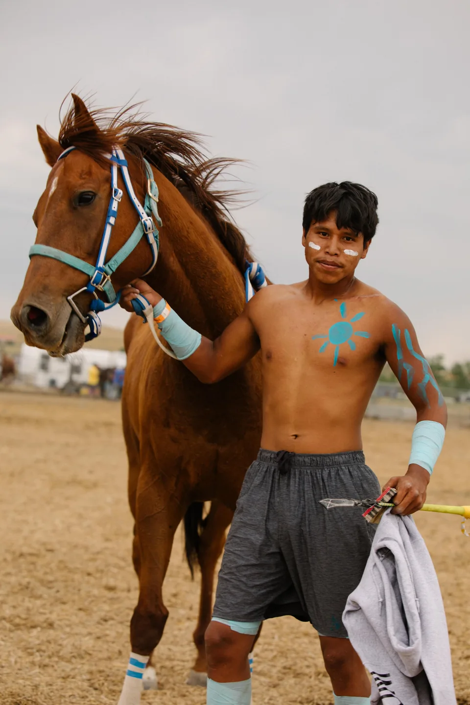A relay rider with turquoise paint on their chest stands beside a brown horse.