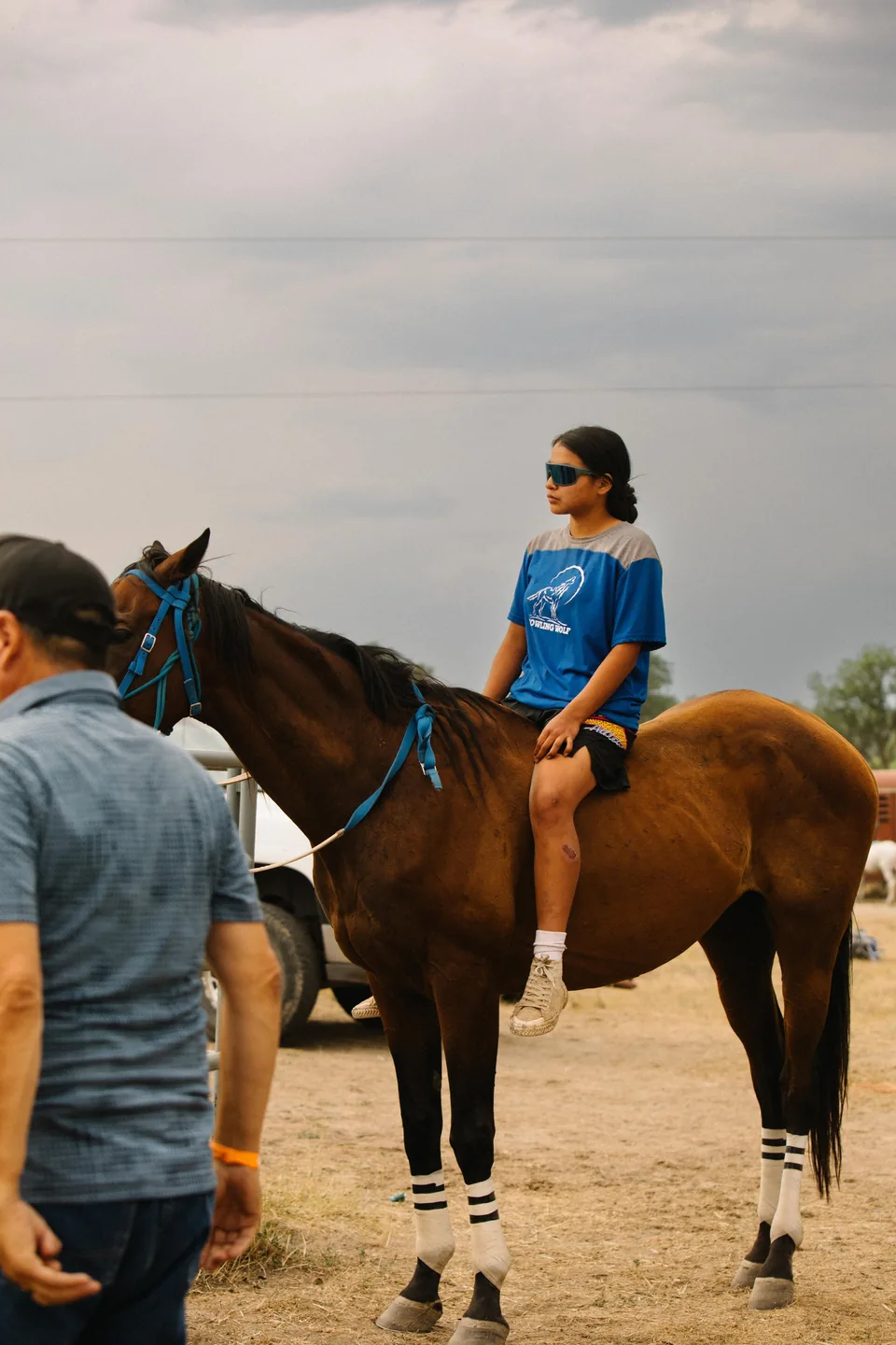 A rider in a blue shirt waits on a brown horse beside a team member.