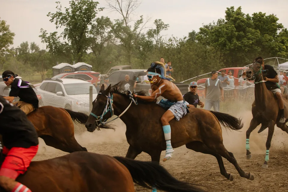 A rider in a blue face covering races past horse holders and parked vehicles.