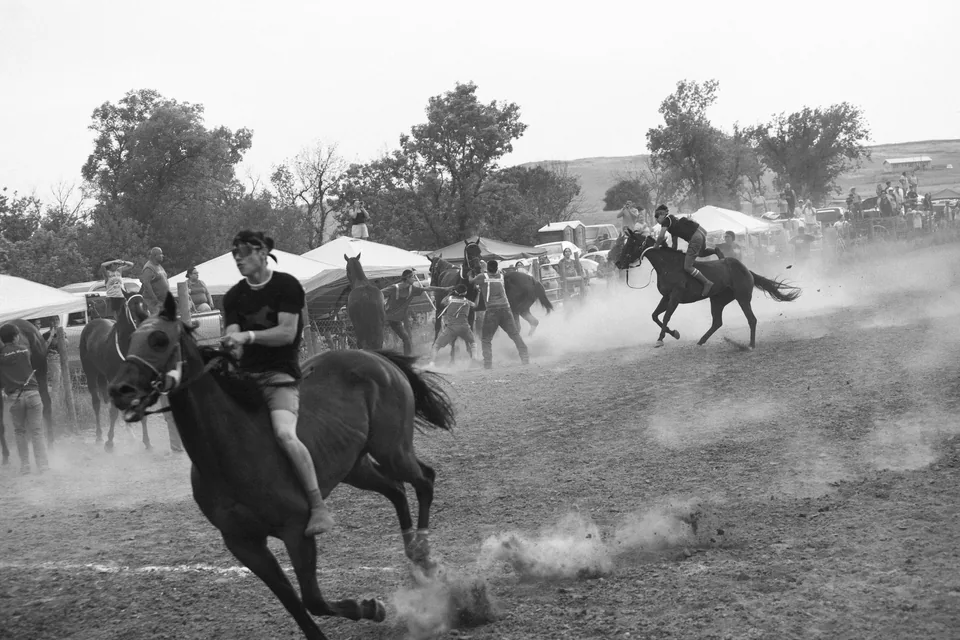 Black-and-white photograph of relay riders moving through dust near the spectators.