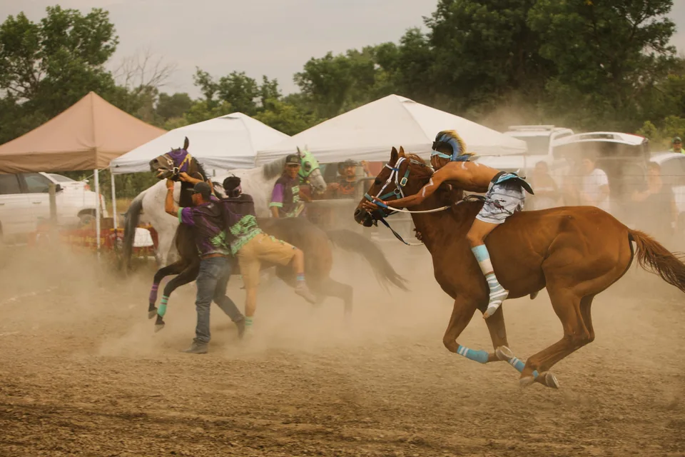 A rider leans low over a brown horse as dust rises along the track.