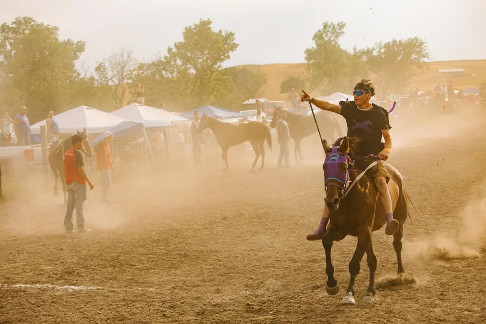 A rider raises the reins while turning a horse through a sunlit cloud of dust.