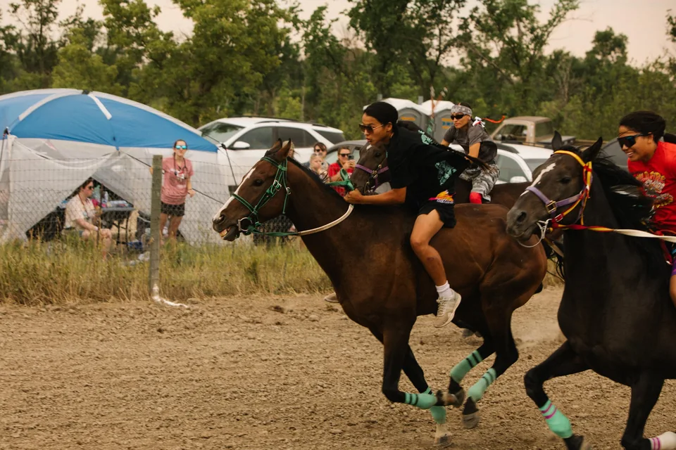 Two bareback riders race past spectators and tents at Pine Ridge.