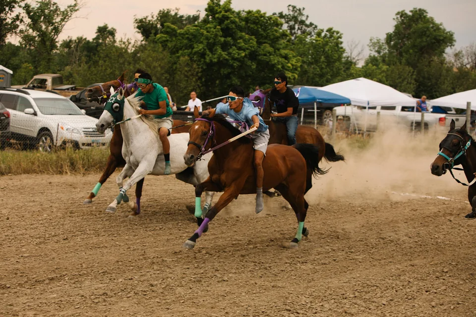 A rider leans forward on a brown horse while racing alongside a white horse.