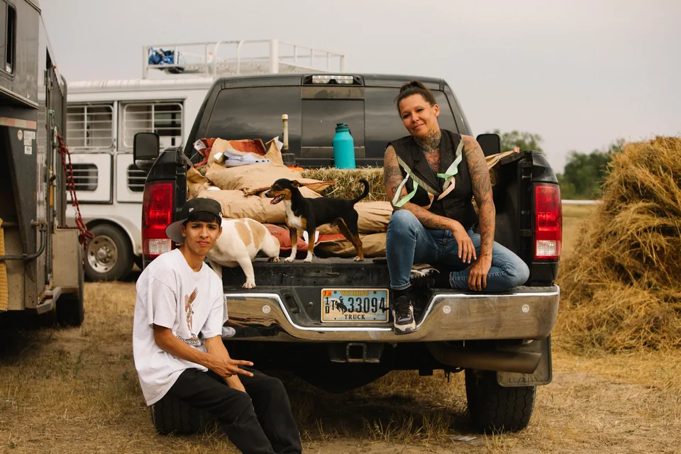 Two people rest on the open tailgate of a pickup beside a horse trailer.