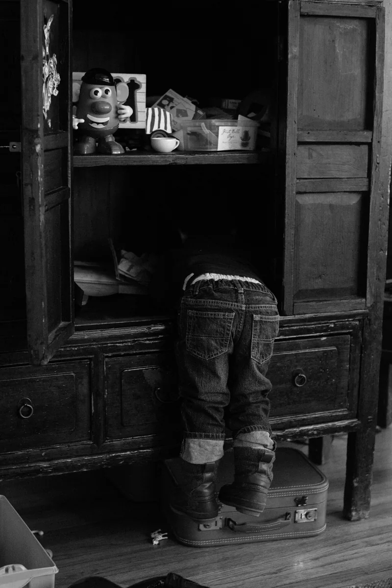A child climbing into an open wooden cabinet, photographed in black and white.