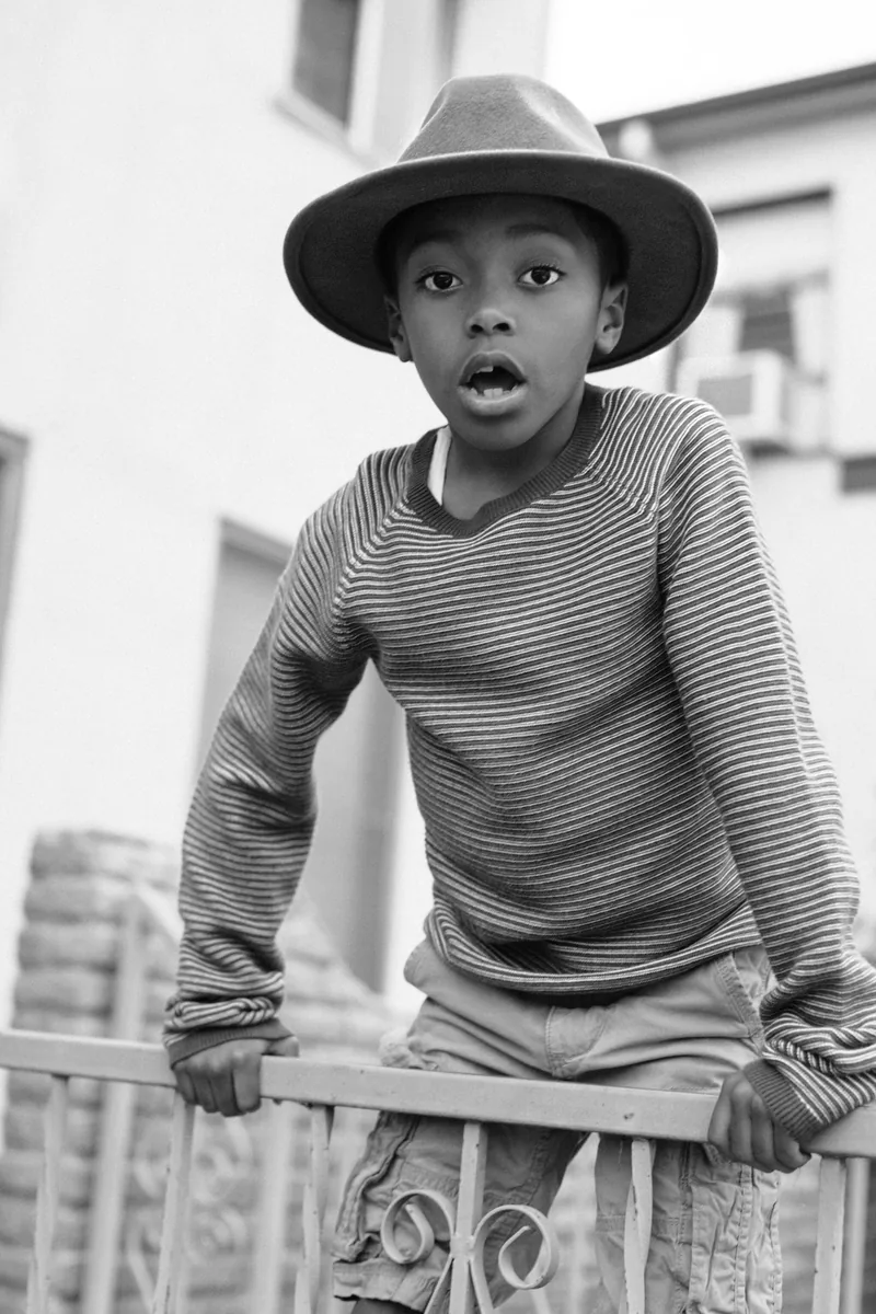 Black-and-white portrait of a child in a wide-brimmed hat leaning over a porch railing.