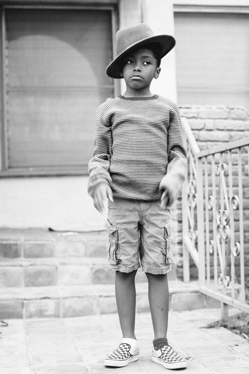 Black-and-white portrait of a child wearing a tilted hat, striped shirt and shorts outside a house.