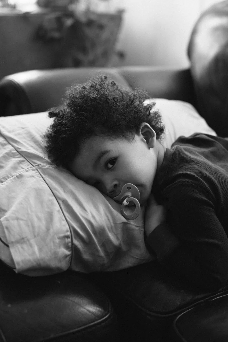 A child resting on a sofa cushion with a pacifier, photographed in black and white.