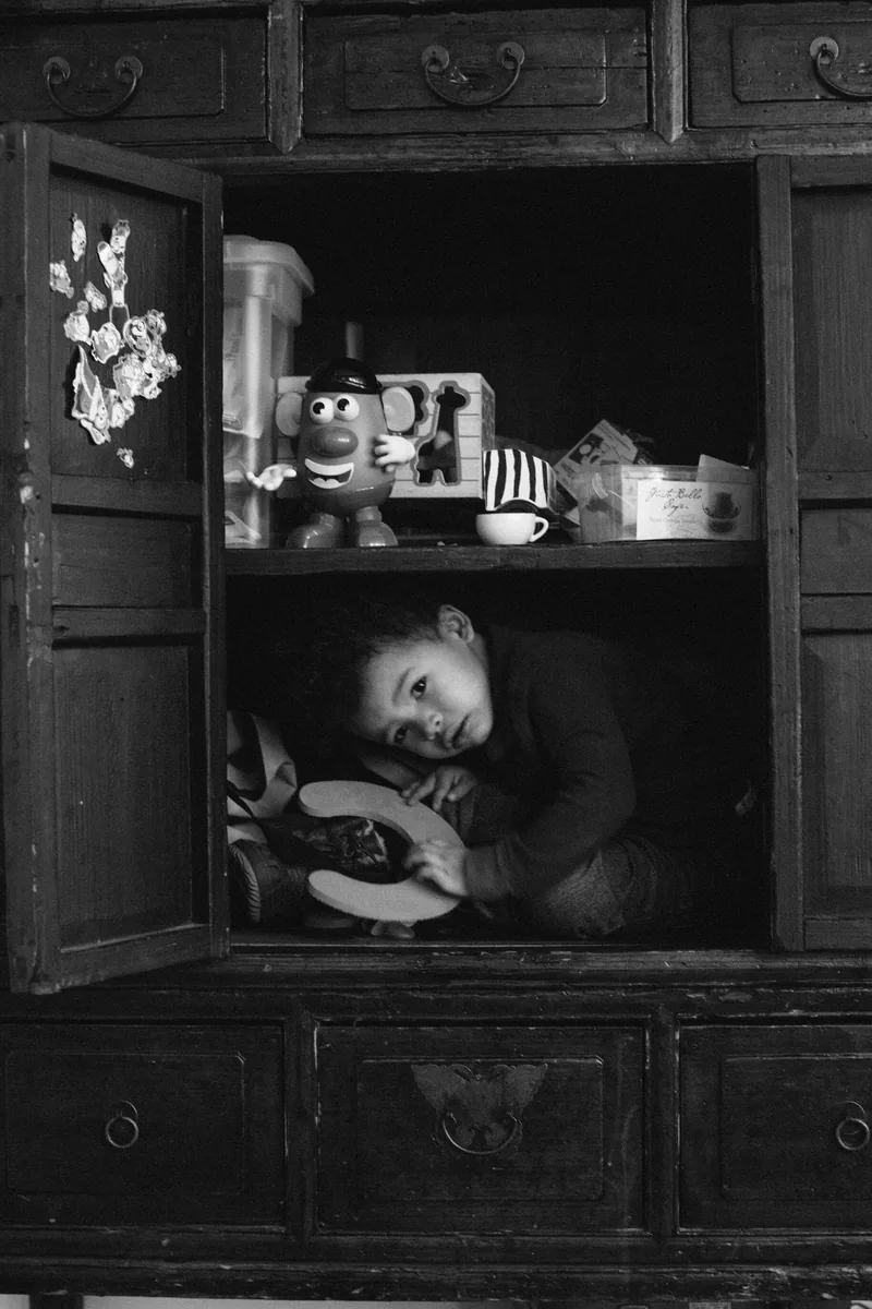 A child curled up inside a wooden cabinet beneath a shelf of cups, in black and white.