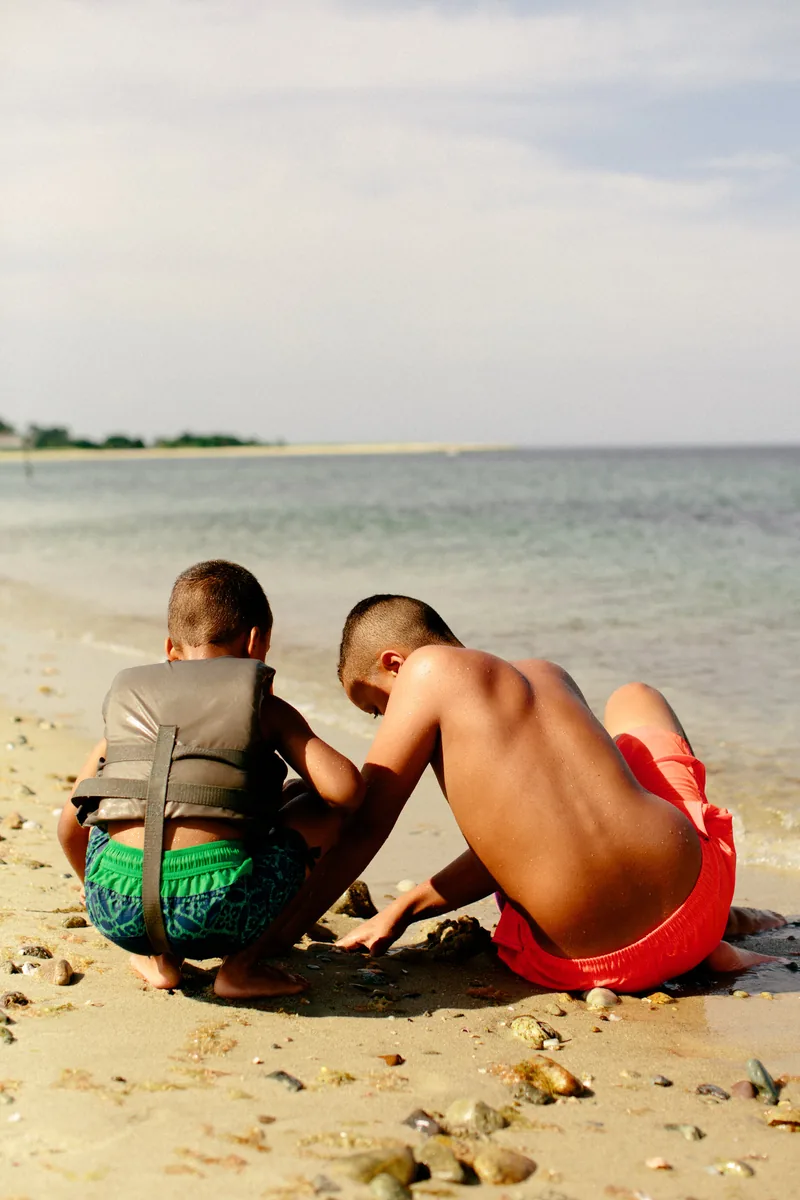Two children crouching together on a beach beside calm blue water.