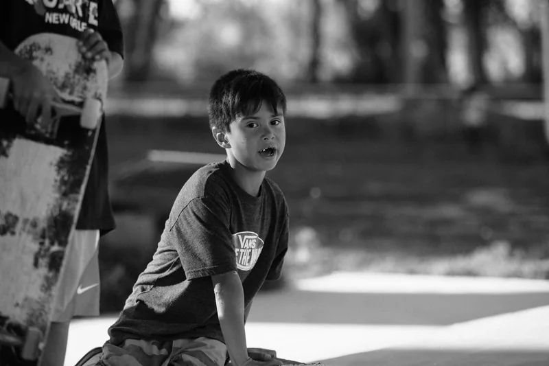 Black-and-white portrait of a child beside an upright skateboard at a skate park.