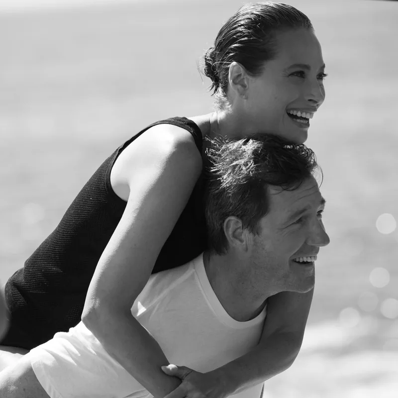 Black-and-white photograph of a smiling model riding piggyback near the water.