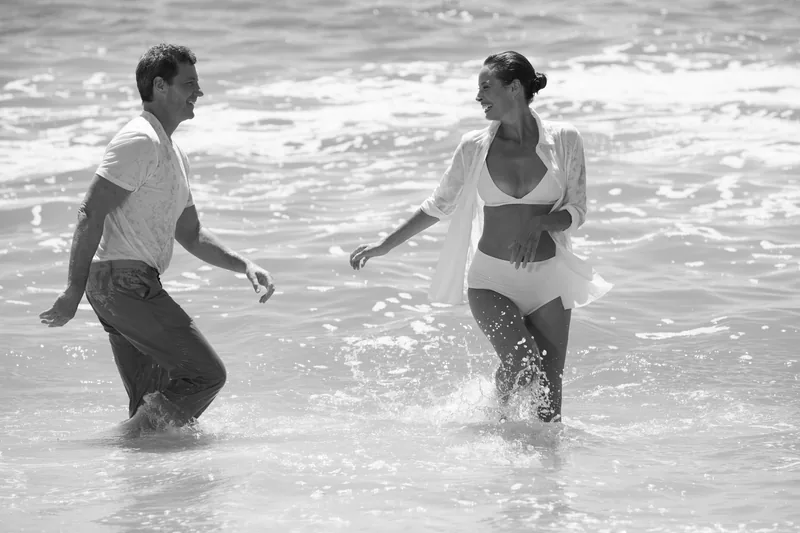Two models walking toward one another through the surf in black and white.