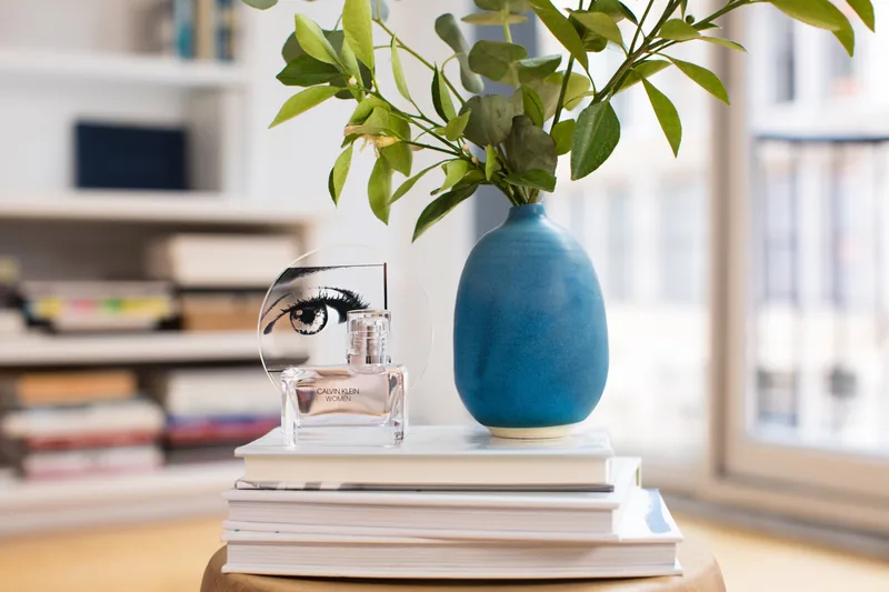 A Calvin Klein Women perfume bottle displayed on books beside a blue vase of leafy branches.