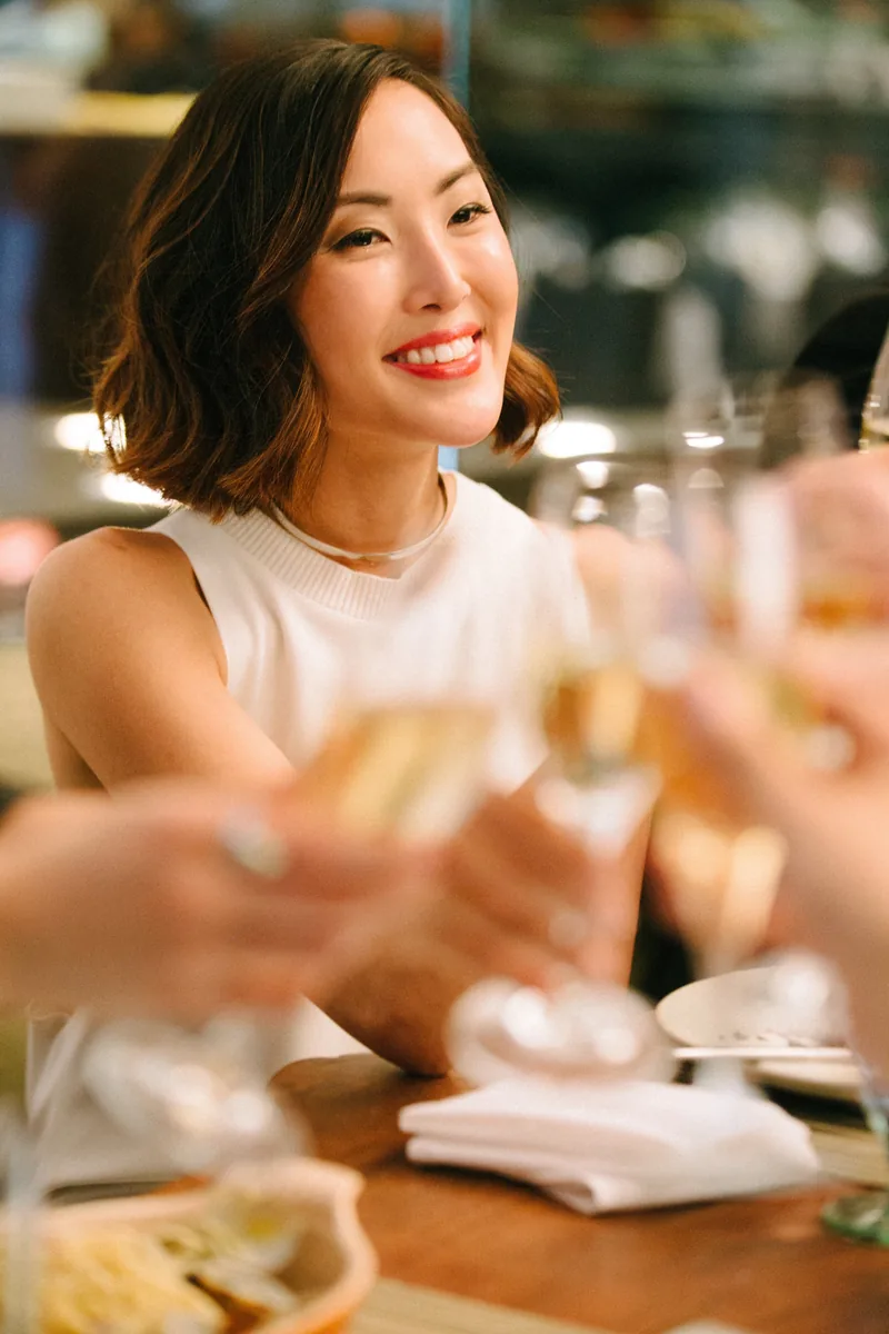 A smiling woman at an outdoor table as glasses are raised for a toast.