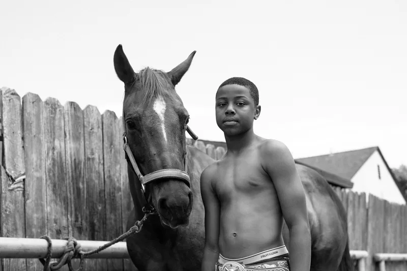 Black-and-white portrait of a young person standing beside a horse at a wooden fence.