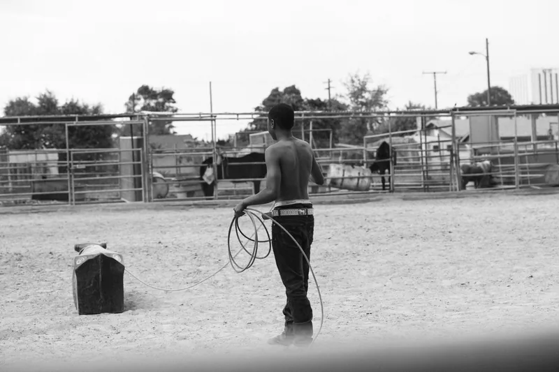 A young person holding a coiled lead rope in a dusty horse enclosure, in black and white.