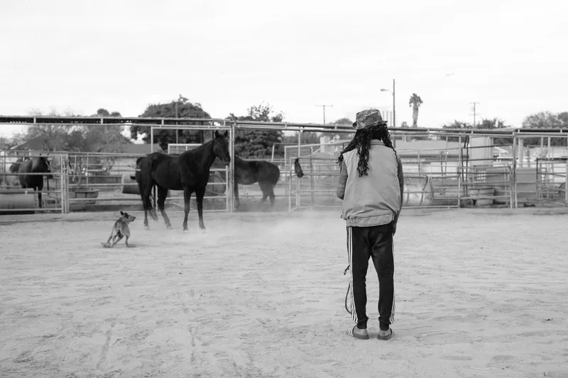 A person walking through a dusty paddock with horses behind them, in black and white.