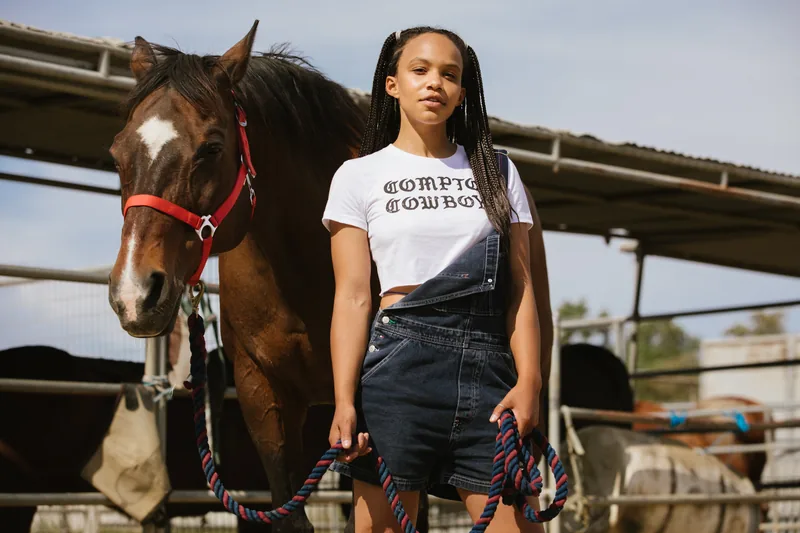 A woman in a Compton Cowboys shirt standing beside a horse with a red halter.