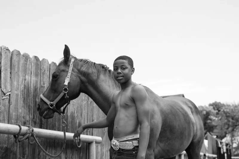Black-and-white portrait of a young person beside a horse, with a large belt buckle at the waist.