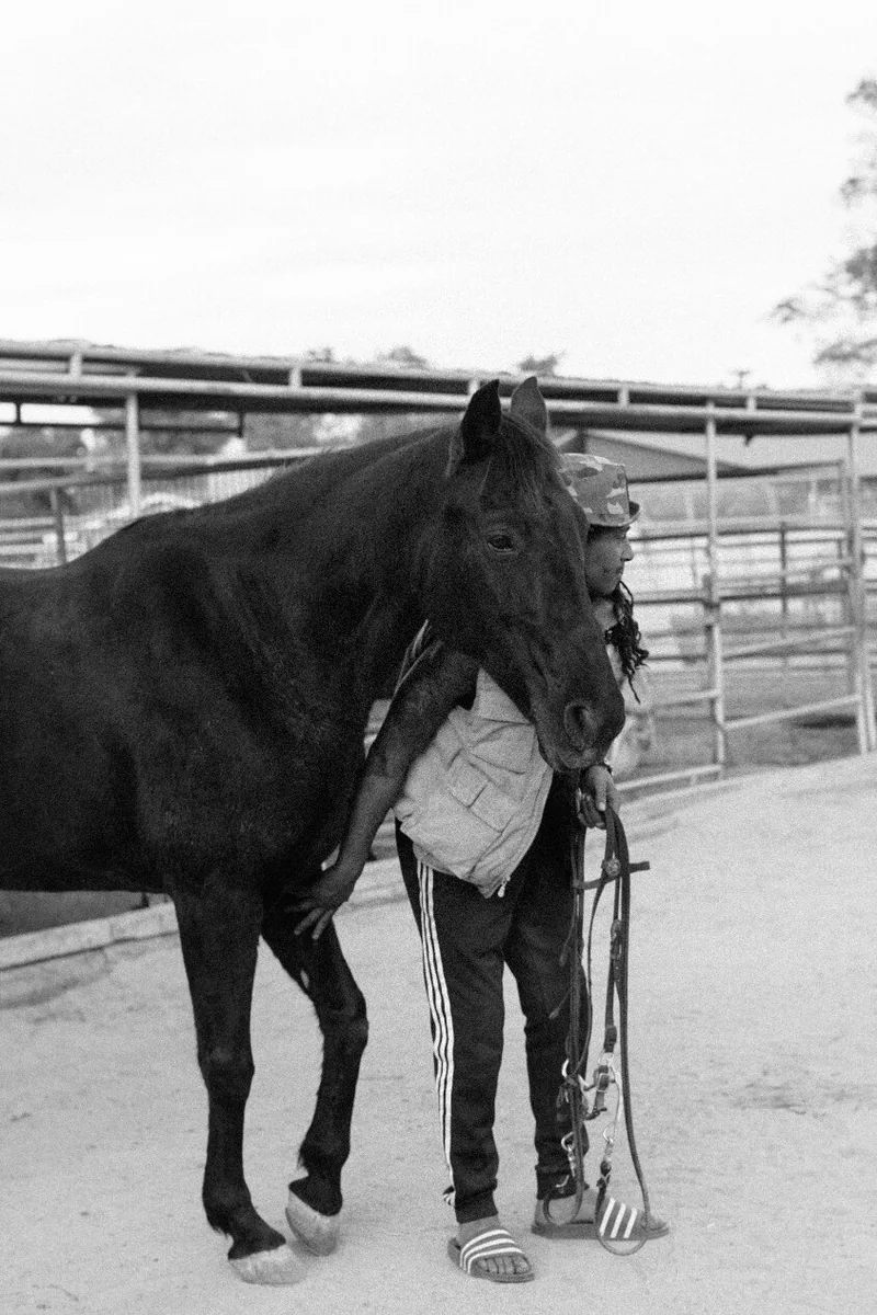 Black-and-white portrait of a person standing close beside a horse in an enclosure.