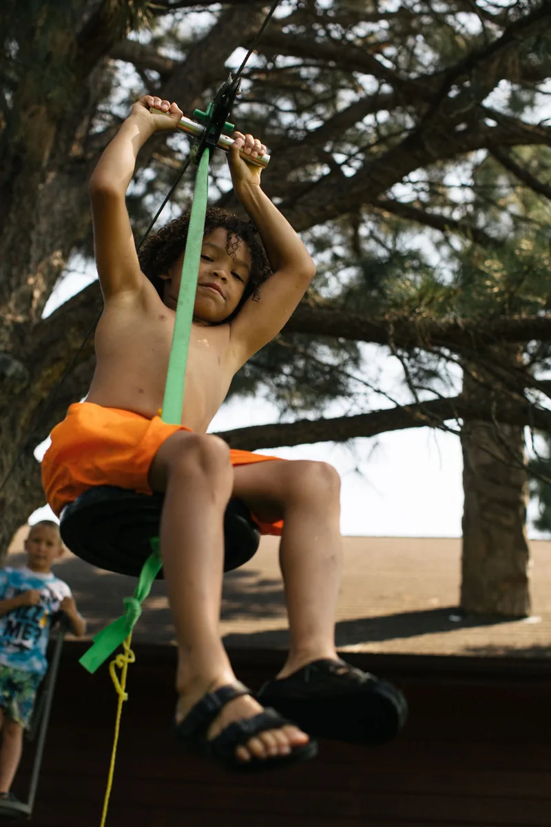 A child in orange shorts holding a rope beneath a spreading tree.