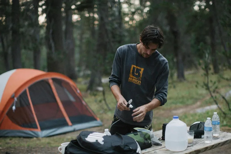 A camper preparing items on a picnic table beside a tent in the woods.