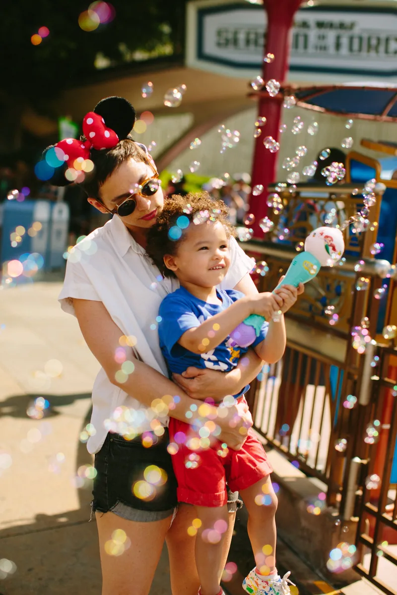 An adult holding a child amid floating bubbles at an outdoor attraction.