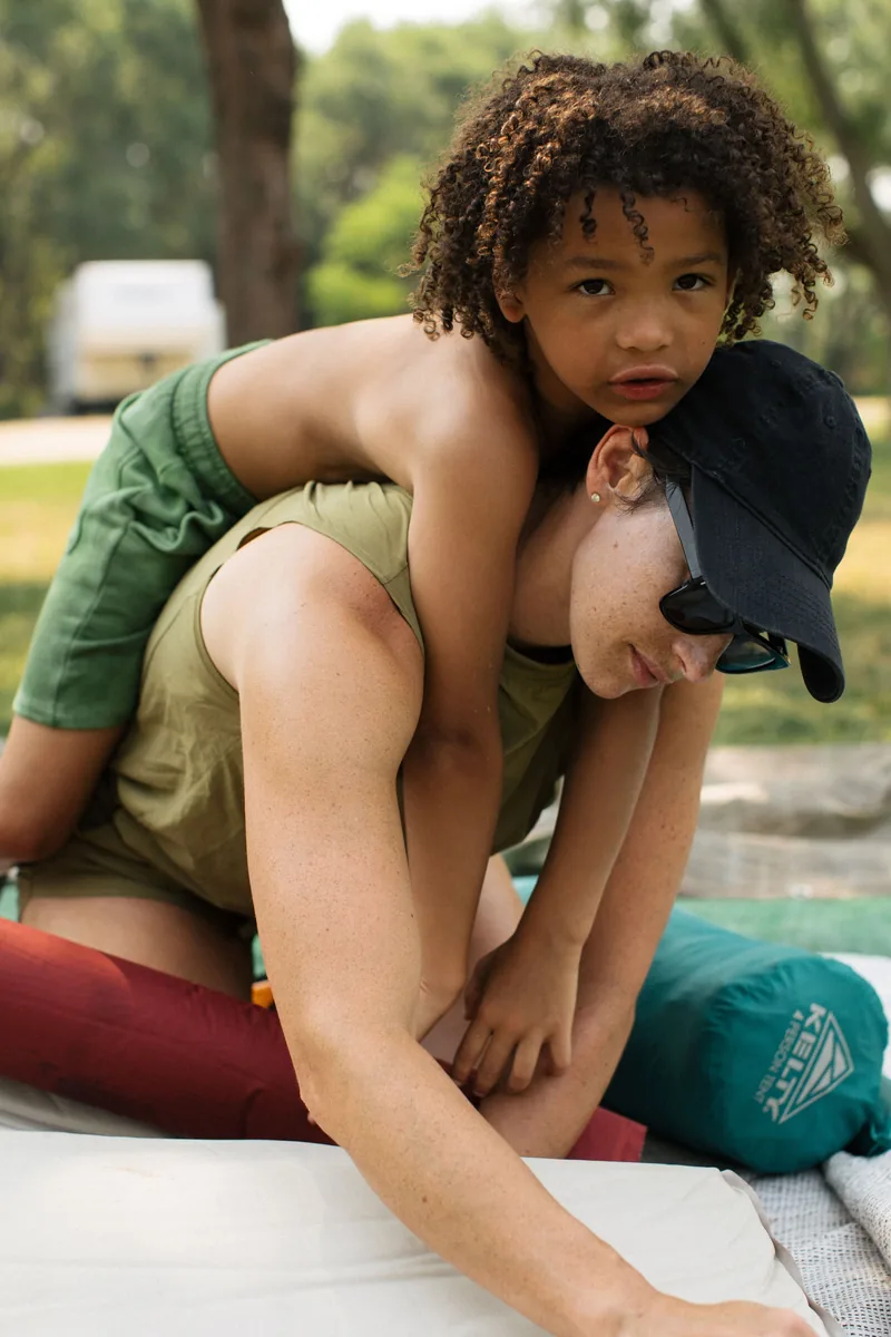 A child leaning over the shoulders of an adult seated outdoors.