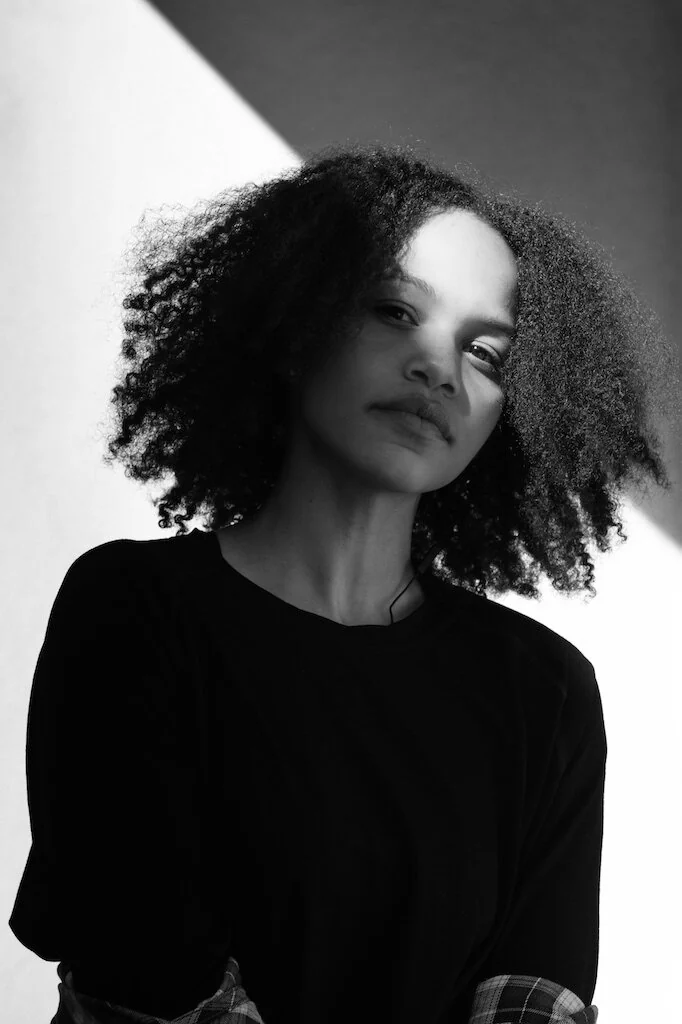 Black-and-white portrait of a woman with curly hair standing in angular window light.