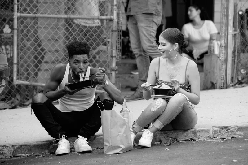 Two people eating from takeaway containers on a curb, photographed in black and white.