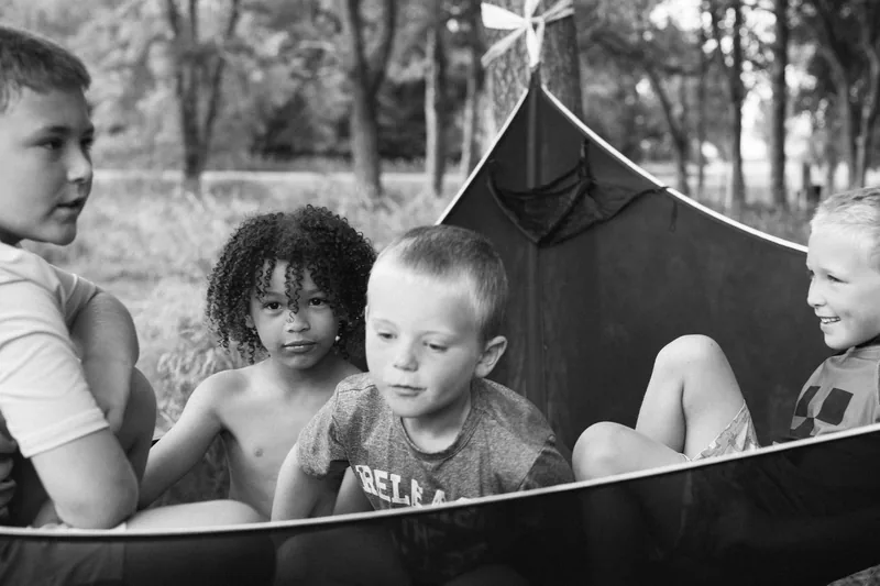 Children gathered in a hammock at a wooded campsite, photographed in black and white.