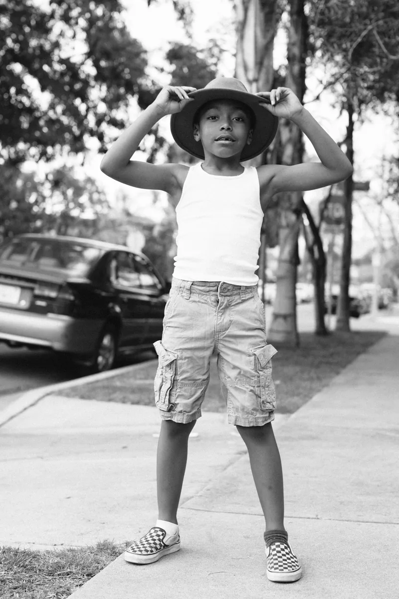 A child holding the brim of a wide hat on a sidewalk, in black and white.