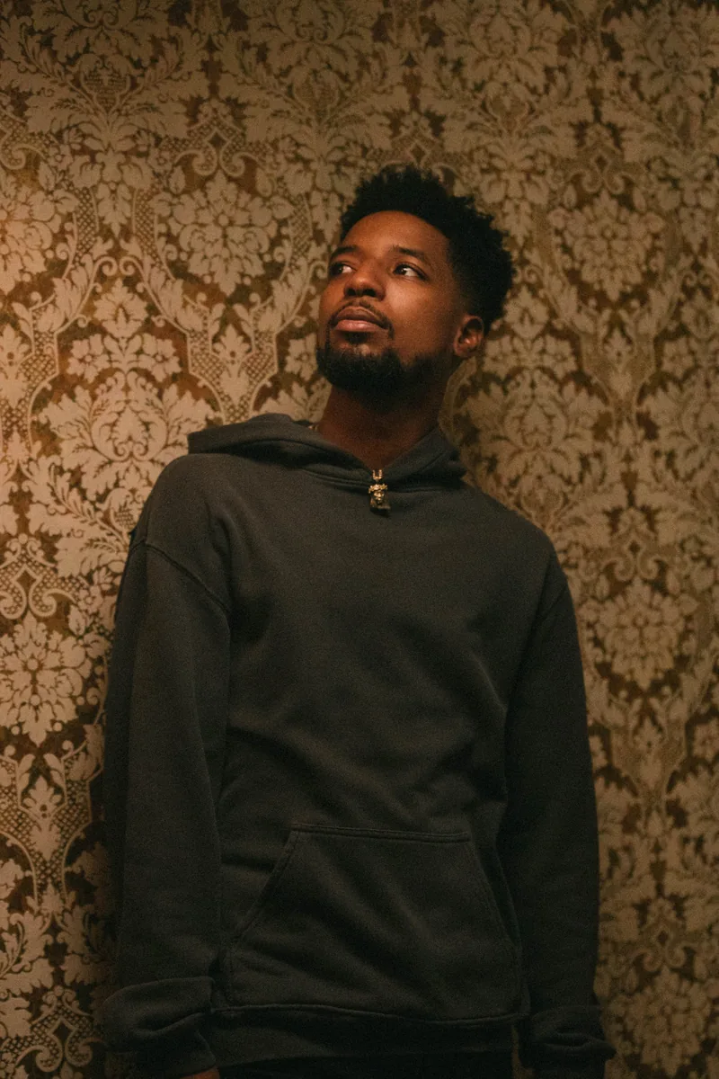 A man in a dark jacket standing against patterned wallpaper.