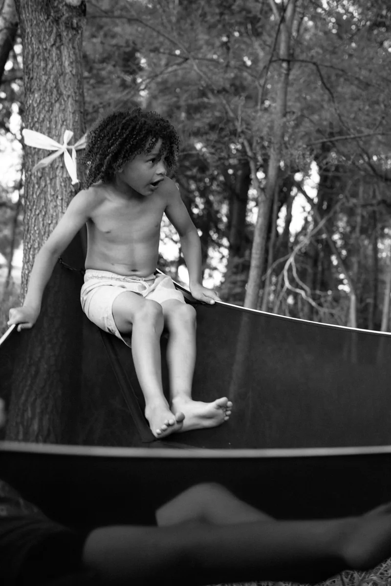 Black-and-white photograph of a child sitting on the edge of a hammock among trees.