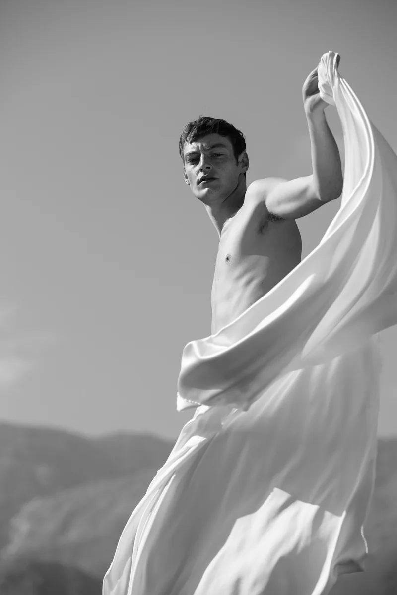 Black-and-white portrait of a model lifting white fabric into the air against a mountain backdrop.