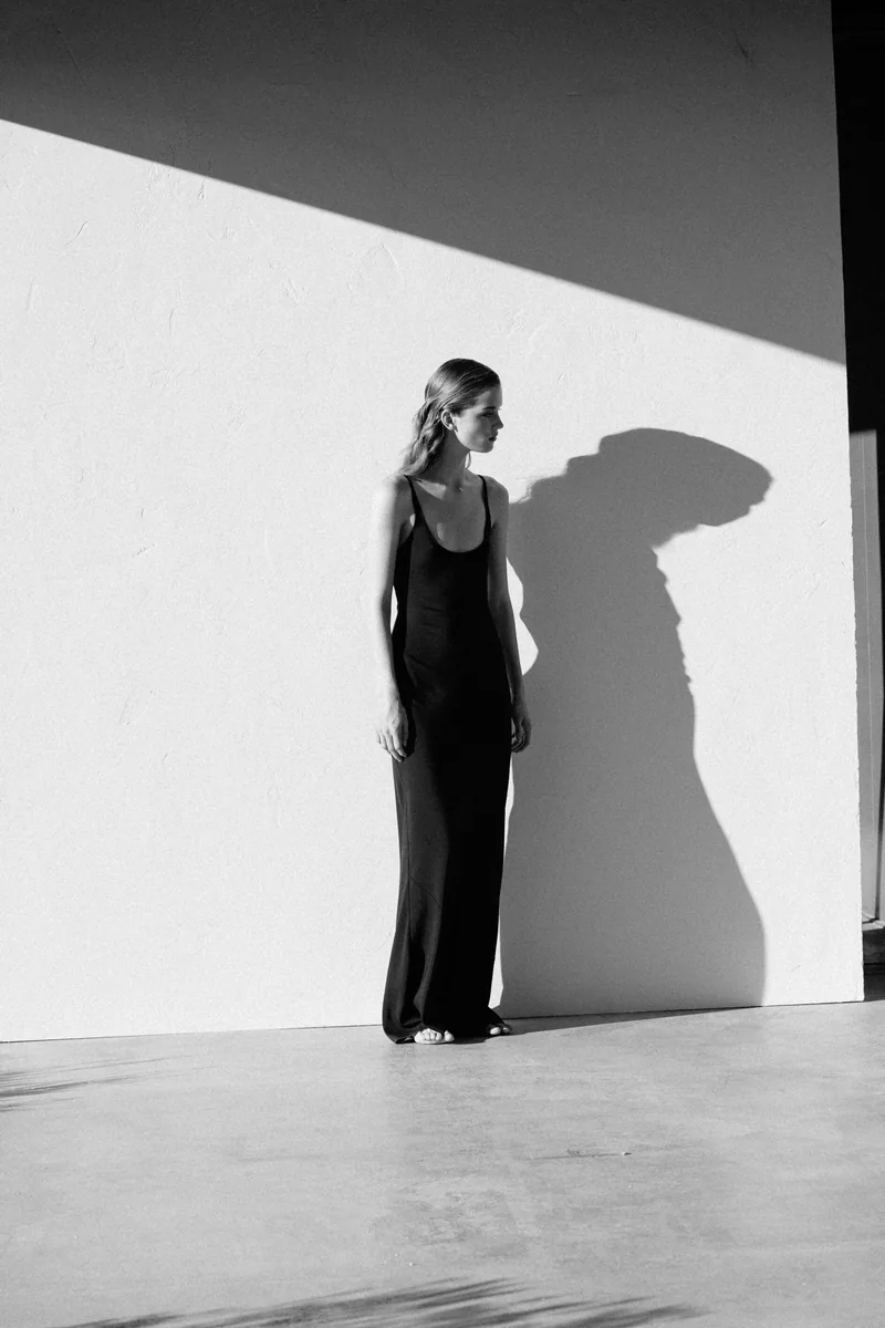 Black-and-white photograph of a model in a long black outfit beside a sharp shadow on a white wall.