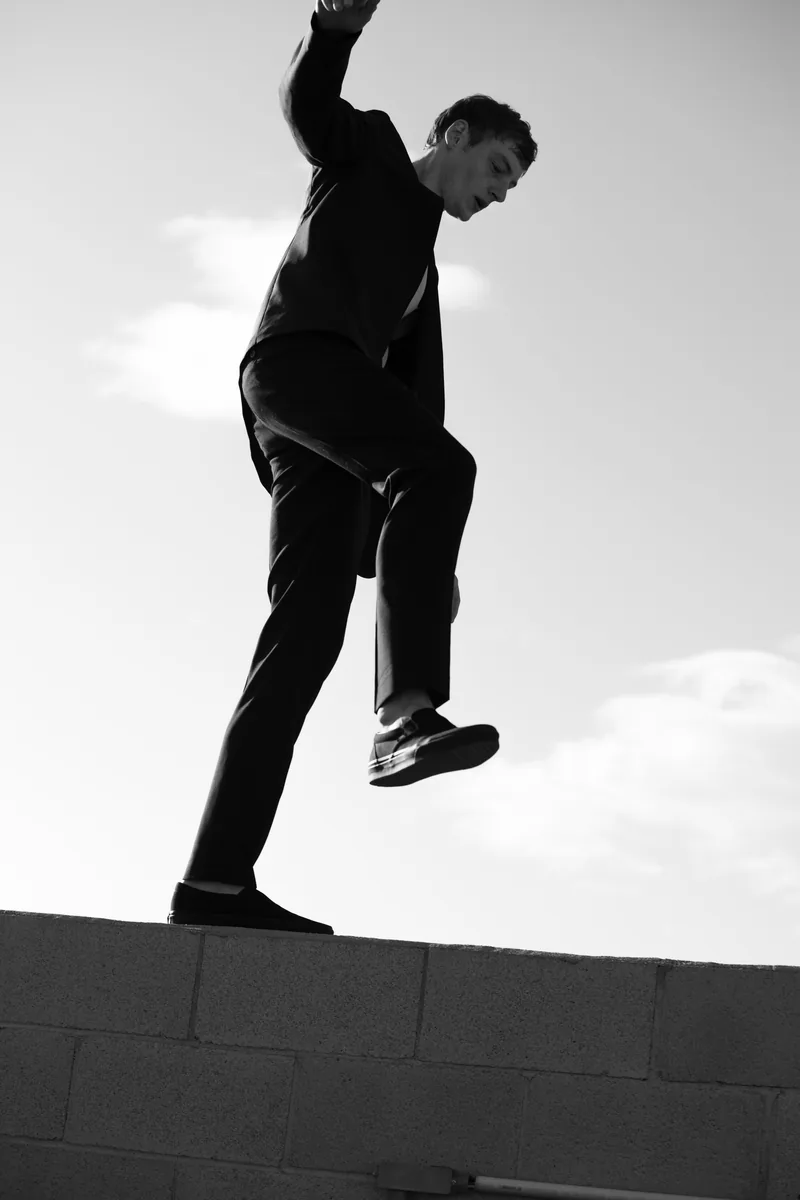 Black-and-white photograph of a model balancing on a low wall with one knee raised.