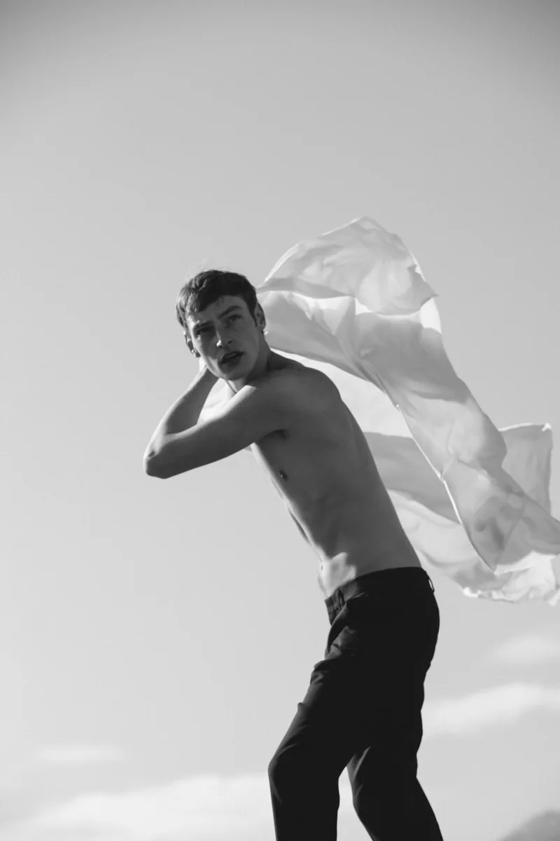 Black-and-white photograph of a model moving with white fabric trailing behind his shoulder.