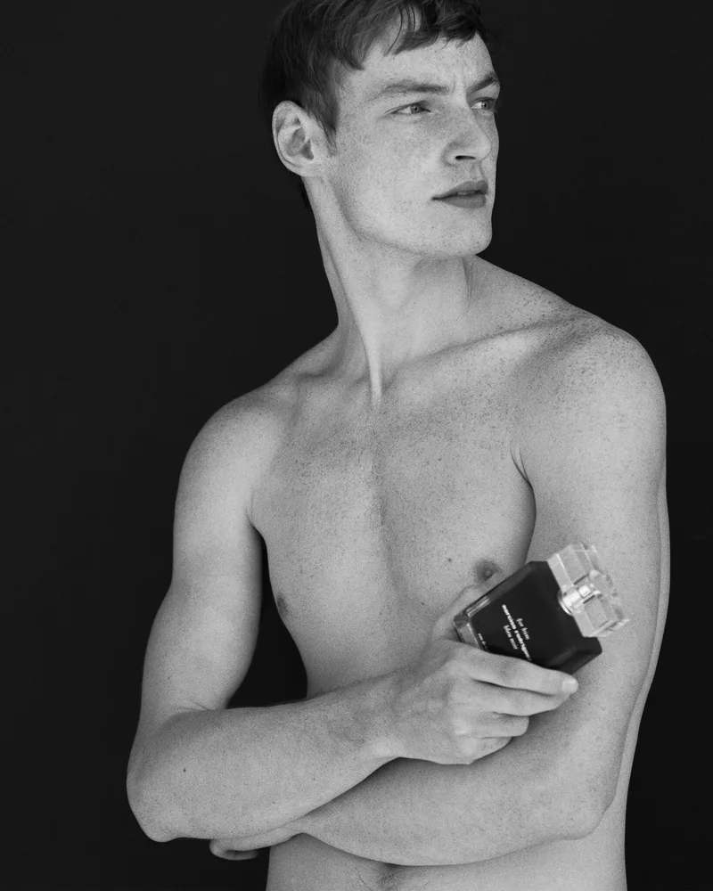 Black-and-white portrait of a model holding a dark perfume bottle against his chest.