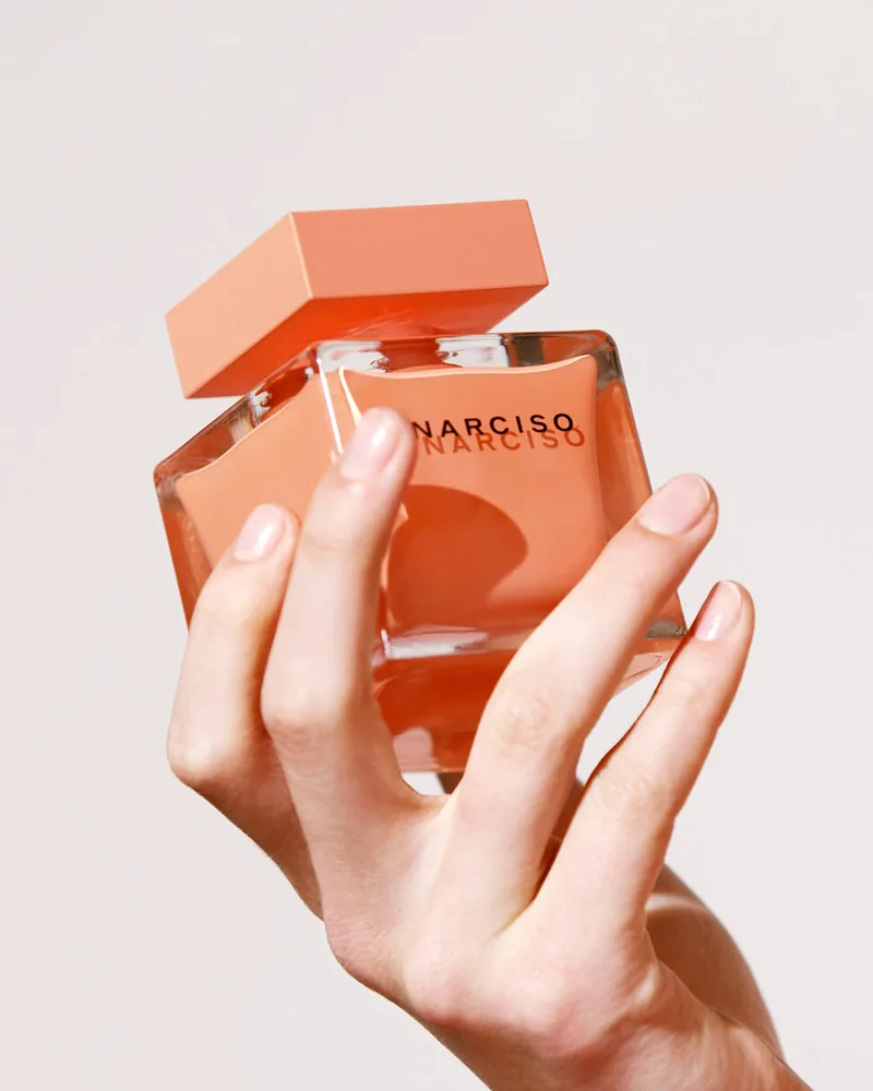 Hands holding a peach-colored Narciso perfume bottle against a pale background.
