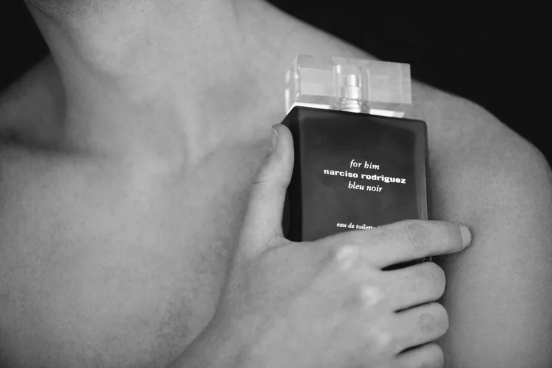 Close-up of a dark Narciso Rodriguez perfume bottle held against a bare shoulder, in black and white.
