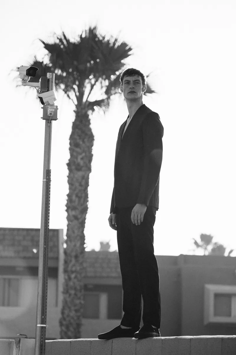 A model in a dark suit standing on a low wall beside palm trees, in black and white.