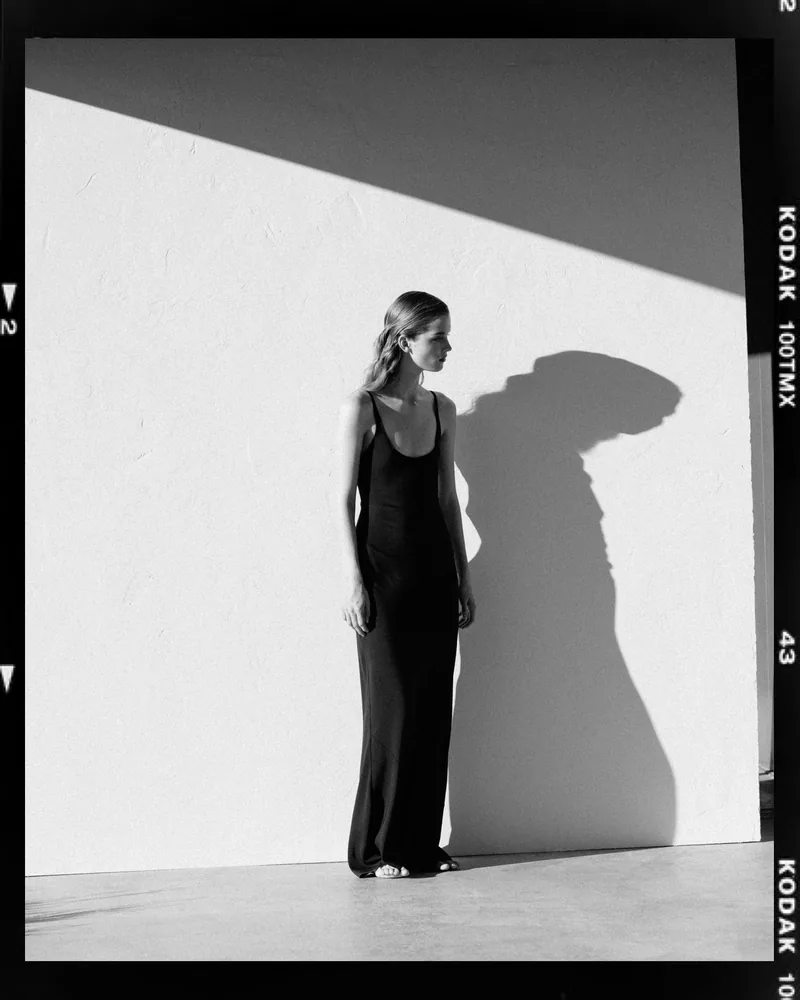 Black-and-white photograph of a model beside her shadow on a white wall, framed by a negative border.