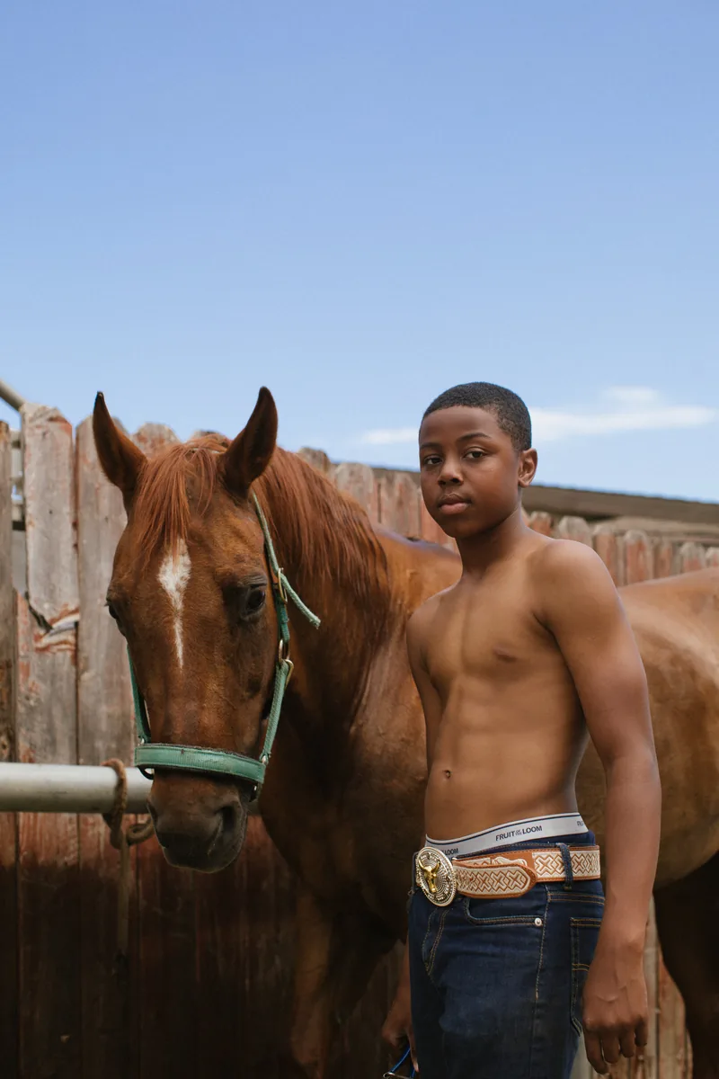 A young person standing beside a brown horse at a wooden fence beneath a clear blue sky.