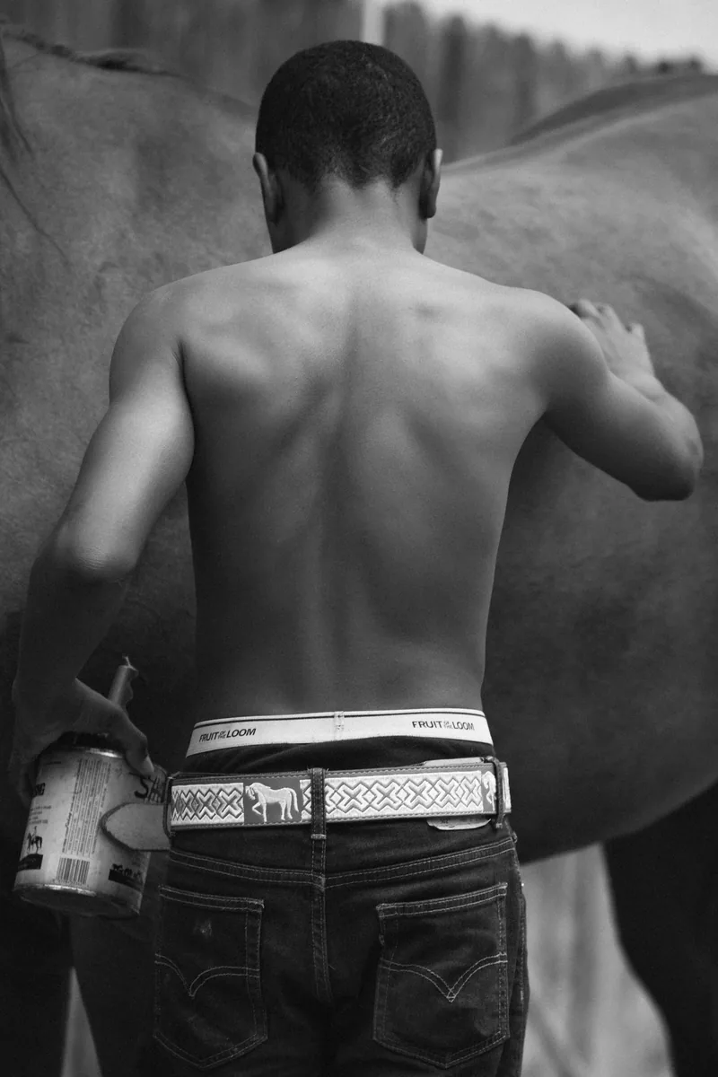 Black-and-white photograph from behind of a young person tending to a horse.